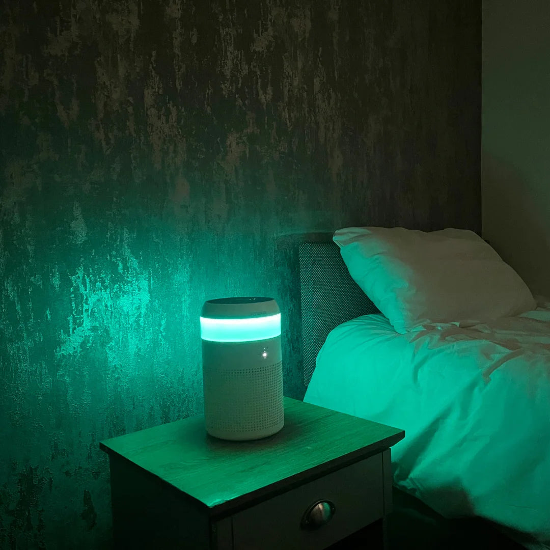 A photo showing the turquoise colour of the LED light inside a Senelux Demi Air Purifier with a bed in the background