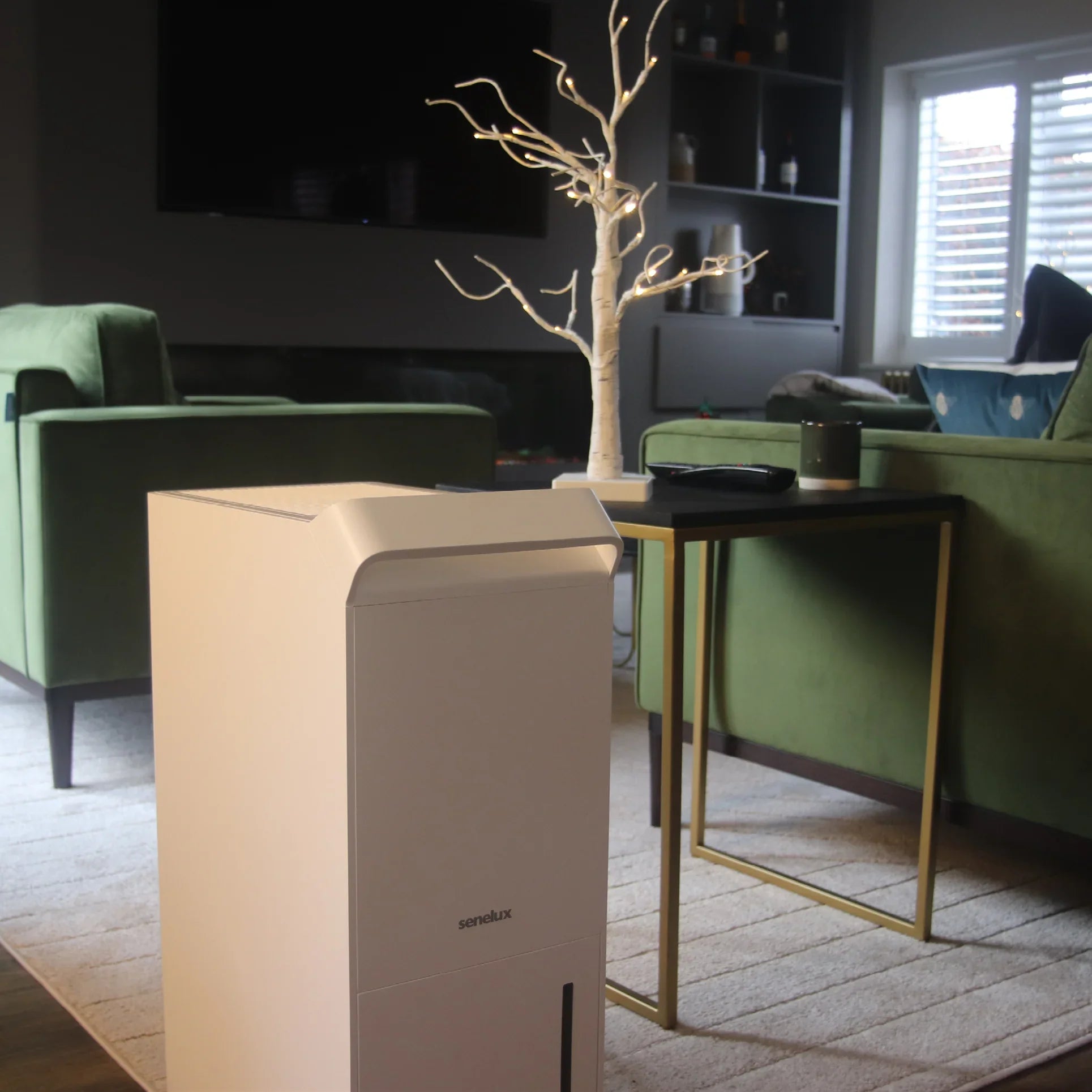A photo of the Senelux 25l/day dehumidifier standing in a room with a green sofa behind it and a stylish Christmas tree on a table to the side 