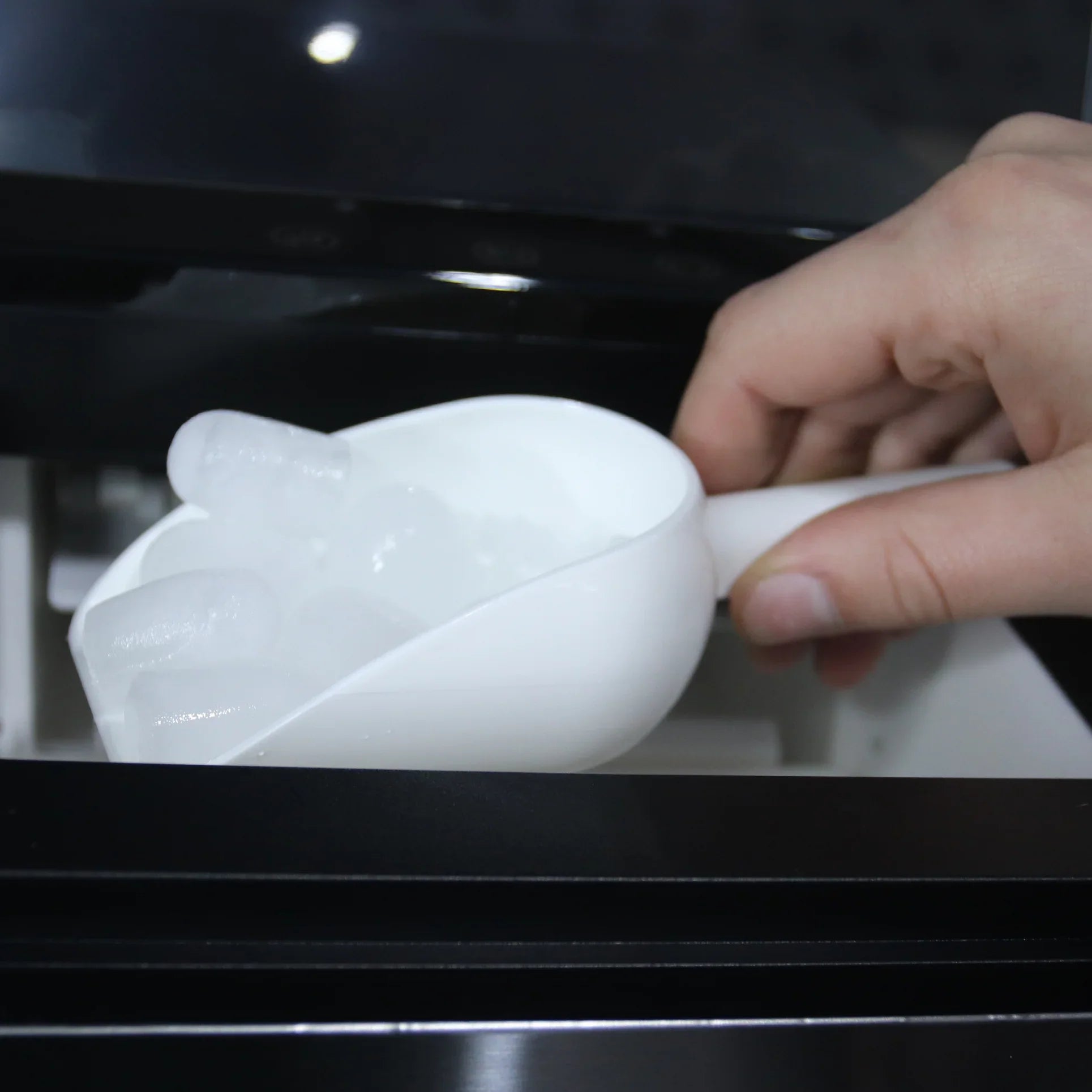 A hand using a clean white scoop to collect ice from a Senelux Compact Ice Maker.
