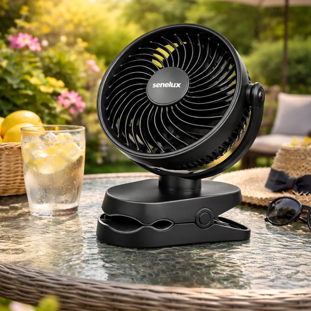 Black fan with 'senelux' branding on a table outdoors, surrounded by a glass of lemonade and a hat.